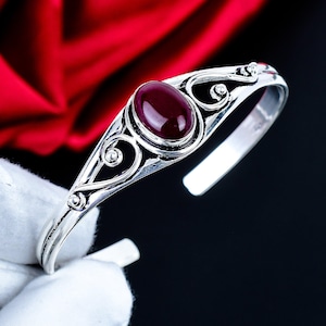 Puede incluir: Un brazalete de plata con una piedra preciosa ovalada de color rojo intenso en el centro. La banda de metal muestra intrincados patrones de volutas caladas y detalles de filigrana a ambos lados del engaste, creando un acabado pulido.