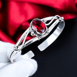 Può includere: Un bracciale a polsino in argento con una gemma ovale rosso intenso. Il bracciale ha un design aperto con decorazioni a volute e piccoli accenti di perline. La gemma è il punto focale, incastonata in una montatura d'argento dettagliata. Lo sfondo è un tessuto rosso sfocato.