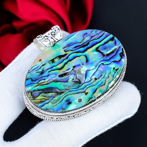 Könnte beinhalten: Ovaler Anhänger mit einer schillernden Abalone-Muschel, die Blau-, Grün- und Lilatöne aufweist. Die Muschel ist in einen detaillierten Silberrahmen gefasst. Der Anhänger wird vor einem weißen Hintergrund präsentiert, um seine natürliche Schönheit hervorzuheben.