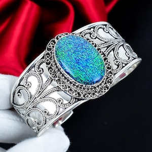 Könnte beinhalten: Ein silbernes Armband mit einem ovalen Mittelstein in strukturierter, schillernder blau-grüner Optik. Das Metallband zeigt komplizierte, durchbrochene Schnörkelmuster und einen Perlenrand, der den zentralen Edelstein umgibt.