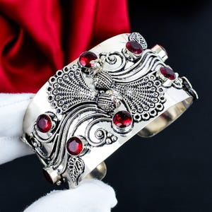 Può includere: Un bracciale a fascia color argento con un motivo a pavone in rilievo, dettagliati disegni di piume e linee sinuose. Il design è arricchito da diverse pietre rotonde sfaccettate di colore rosso intenso incastonate nel metallo.