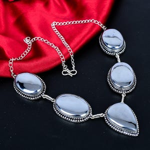 Puede incluir: Un collar plateado con piedras grises grandes, ovaladas y en forma de lágrima. Las piedras están engastadas en un marco plateado con cuentas y conectadas por una cadena plateada. El collar se exhibe sobre un fondo de satén rojo.