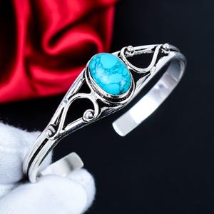 May include: Silver cuff bracelet with an oval turquoise stone. The openwork design features decorative swirls and small ball accents. The turquoise stone is a bright blue-green colour. The bracelet is shown against a black background.
