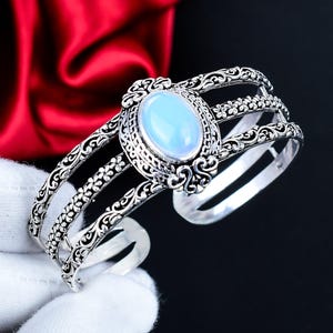 Puede incluir: Brazalete de puño plateado con una piedra preciosa ovalada de color azul pálido en el centro. El brazalete tiene un diseño abierto con intrincados patrones florales y de volutas. La piedra preciosa está engastada en un bisel detallado.