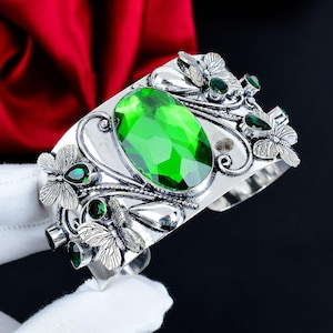 Peut inclure: Un bracelet manchette en argent orné d'une grande pierre précieuse ovale d'un vert éclatant au centre. Le métal présente des motifs de papillons détaillés et des petites pierres vertes en forme de goutte sur la surface.