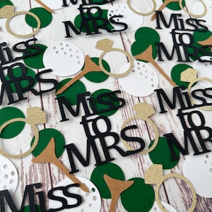 May include: Confetti with a wedding theme, featuring the text "Miss to Mrs" in black, gold glitter rings, golf tees, and green and white circles. The confetti is scattered on a light wood grain surface.
