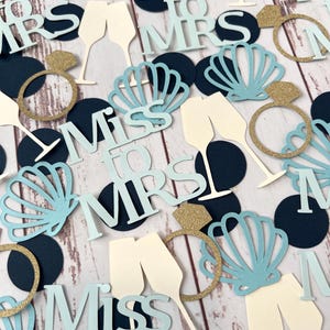 Op de afbeelding: Bruiloft-thema confetti met champagneglazen, verlovingsringen en schelpvormen. De confetti bevat de zinnen "Miss to Mrs" in lichtblauw, goudglitter en marineblauw. De achtergrond is een lichte houtnerf.