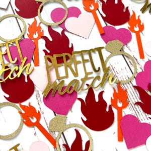 May include: Gold glitter confetti with the words "perfect match" and images of hearts, rings, and flames. The confetti is on a white wooden background.