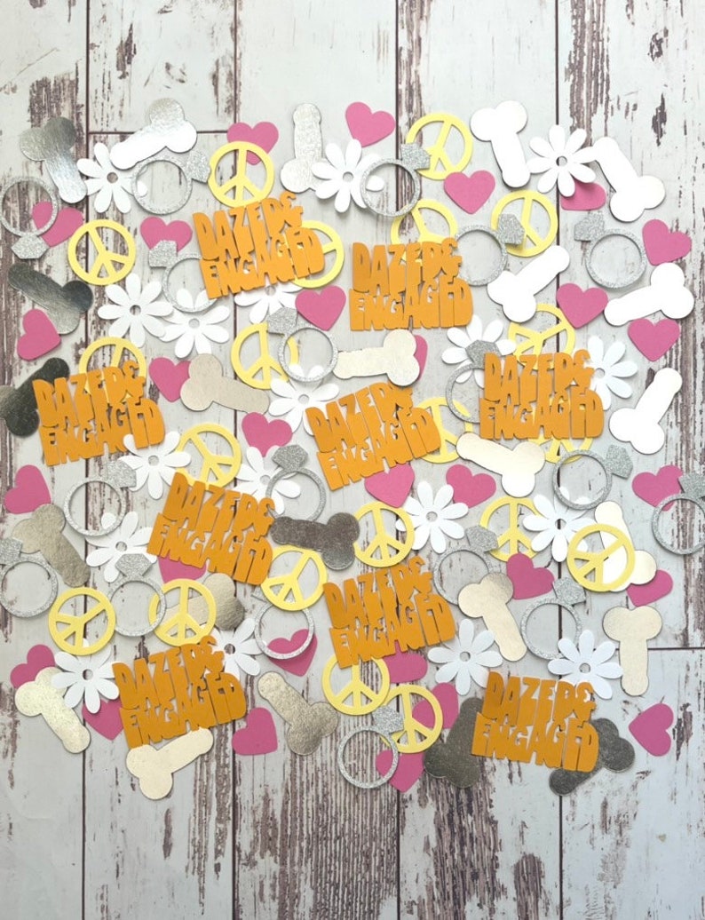 Pode incluir: Uma variedade de confete com sinais de paz, an&eacute;is, cora&ccedil;&otilde;es, flores e o texto "Dazed & Engaged" em glitter dourado. O confete est&aacute; espalhado sobre uma superf&iacute;cie de madeira branca.