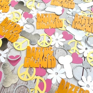 Pode incluir: Confete para um casamento ou uma festa de noivado. O confete tem a forma de sinais de paz, cora&ccedil;&otilde;es, flores e an&eacute;is. O confete &eacute; amarelo, rosa, branco e prateado. O confete tem o texto "Dazed & Engaged".