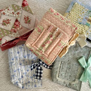 May include: A collection of quilted fabric pouches in various floral and patterned designs. The pouches feature pink, blue, green, and yellow fabrics with ribbon and bow accents. Some pouches have pockets for pens or small items.