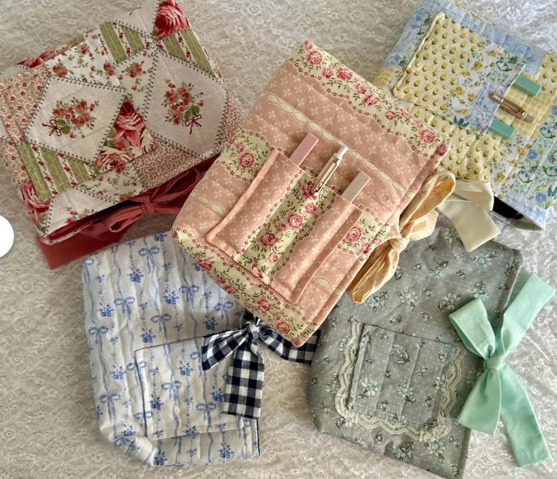 May include: A collection of quilted fabric pouches in various floral and patterned designs. The pouches feature pink, blue, green, and yellow fabrics with ribbon and bow accents. Some pouches have pockets for pens or small items.