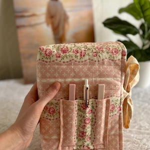 May include: A quilted, floral-patterned book cover in shades of pink and cream. The cover features pockets holding a pen and two lipsticks. A decorative bow is tied on the side. The background includes a painting and a houseplant.