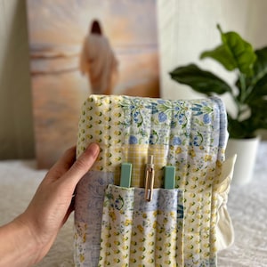 May include: A floral patterned quilted book cover in yellow, blue, and white. It features a pocket containing two pale green pens and a gold pen. A painting and a potted plant are visible in the background.