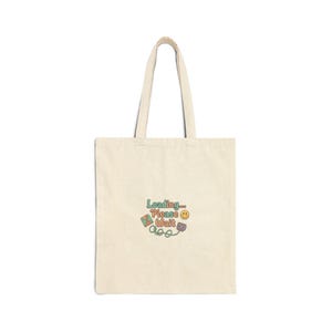May include: Cream-colored tote bag with a retro-style graphic that reads "Loading... Please Wait" in green, orange, and purple. The design includes a smiling face and a game controller. The bag has two long handles.
