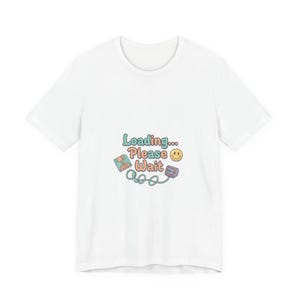 Könnte beinhalten: Weißes T-Shirt mit einem farbenfrohen Aufdruck "Loading... Please Wait" mit einer Diskette, einer Spielkonsole und einem Smiley. Das Design verwendet Pastellfarben.