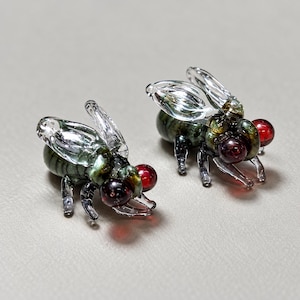 May include: Two small glass insect figurines featuring clear, translucent wings, dark green striped bodies, and prominent, spherical red eyes. Each piece is crafted with detailed legs and antennae, showcasing a glossy finish and intricate glasswork.