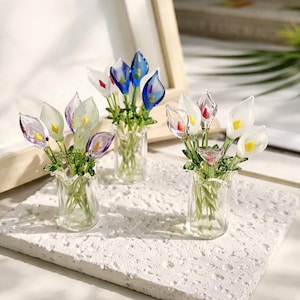 Könnte beinhalten: Miniatur-Calla-Lilien-Bouquets in klaren Vasen. Die Blumen haben durchscheinende Blütenblätter in Weiß, Blau, Lila und Rot, mit gelben Zentren und grünen Stielen. Die Vasen sind klar mit einem gerüschten Rand.