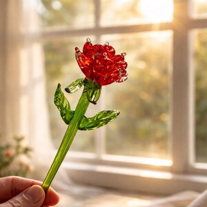May include: A handcrafted glass rose with a vibrant red bloom and green stem and leaves. The delicate petals and leaves are detailed, showcasing the artistry of the glasswork. The rose is held in a hand, with a window and sunlight in the background.