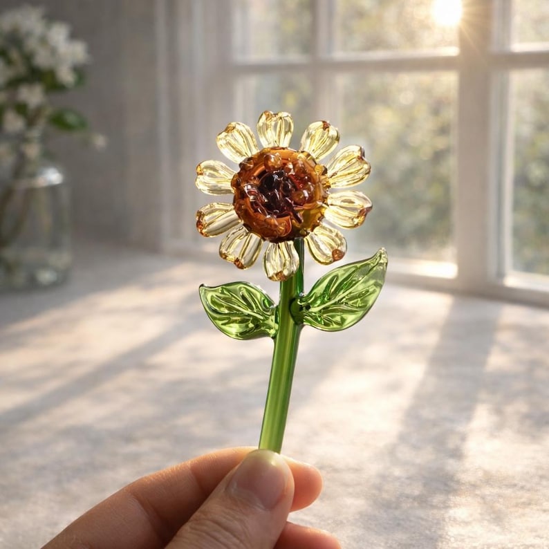 Puede incluir: Un girasol de cristal hecho a mano con un centro marr&oacute;n y p&eacute;talos amarillos transl&uacute;cidos. El tallo y las hojas son verdes. La flor se sostiene en una mano, con una ventana y luz solar en el fondo.