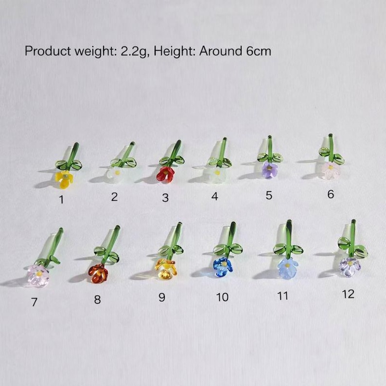 May include: A collection of twelve miniature glass flower sculptures with green stems and leaves. The flowers come in various colors, including yellow, white, red, purple, and blue. Each flower is approximately 2.4 inches tall.