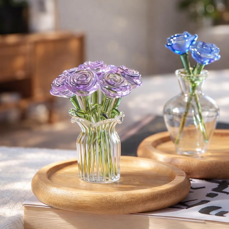 May include: A clear glass vase with a ruffled rim holds a bouquet of purple glass roses. Another small vase with two blue glass roses sits on a wooden tray. The vases are on a wooden surface.
