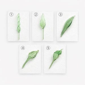 May include: Five pieces of green glass botanical art, each depicting a different stage of a leaf or stem. The pieces are numbered 1 through 5 and are set against a white background. The glass has a delicate, translucent quality.