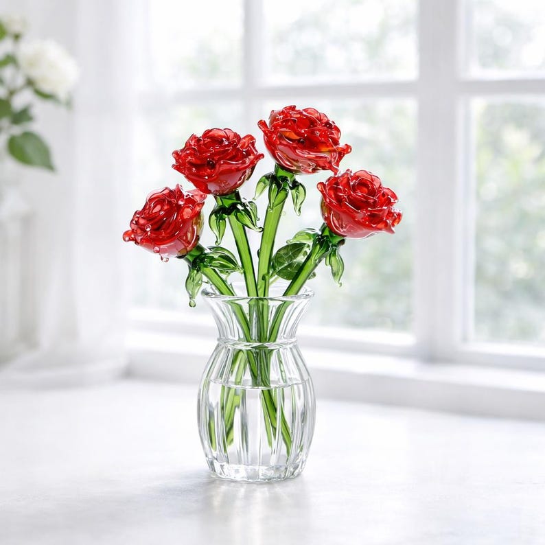 May include: A clear glass vase holding a bouquet of five red glass roses. The roses have green stems and leaves. The vase is sitting on a white surface in front of a window.