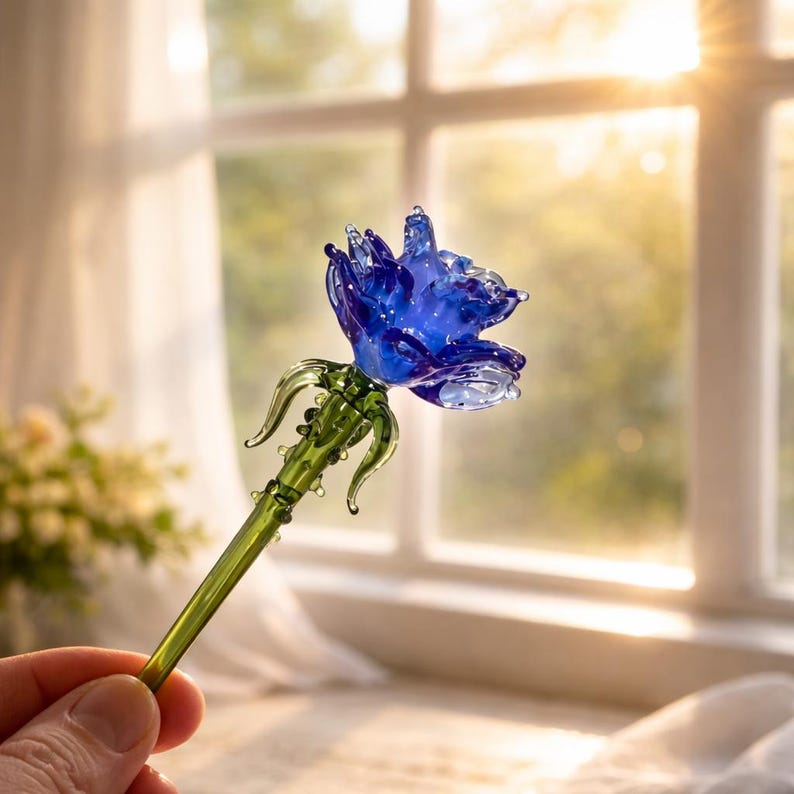 May include: A handcrafted glass rose with a vibrant blue flower and a green stem. The delicate petals of the rose are intricately detailed, and the stem features small, decorative leaves. The glass rose is a decorative item.