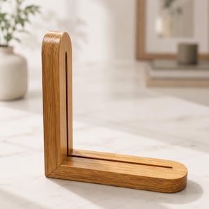 May include: An L-shaped wooden display stand crafted from light-toned oak with a visible wood grain. The stand features a vertical upright piece and a horizontal base, both containing a long, narrow central slot for holding items upright.