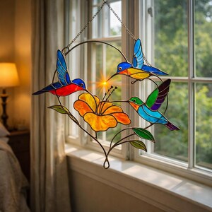 K&ouml;nnte beinhalten: Buntes Glasfensterbild mit drei Kolibris in Blau, Rot und Gr&uuml;n, einer gelben Blume und gr&uuml;nen Bl&auml;ttern. Das Fensterbild hat einen Metallrahmen und eine Kette zum Aufh&auml;ngen. Die Dekoration ist vor einem Fenster angebracht.