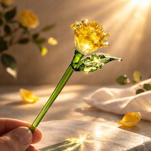 May include: A handcrafted glass rose with a yellow flower head, green stem, and a single leaf. The rose is held in a hand, with a soft background and sunlight shining through the flower. The glass rose is a decorative item.