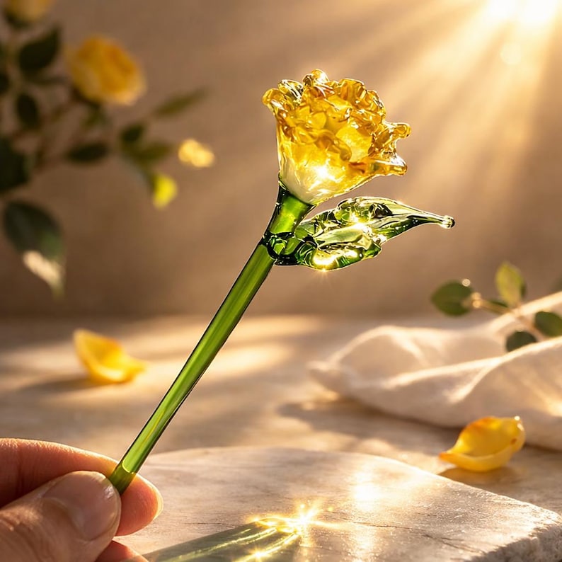 May include: A handcrafted glass rose with a yellow flower head, green stem, and a single leaf. The rose is held in a hand, with a soft background and sunlight shining through the flower. The glass rose is a decorative item.