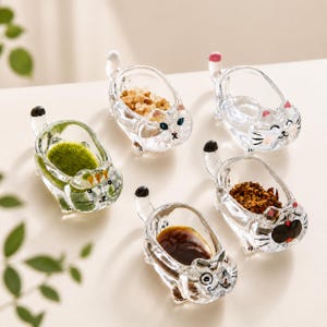 May include: A collection of five clear glass cat-shaped condiment dishes. Each dish is filled with different ingredients, including salt, spices, and sauces. The dishes have black and pink accents and are arranged on a light-colored surface.