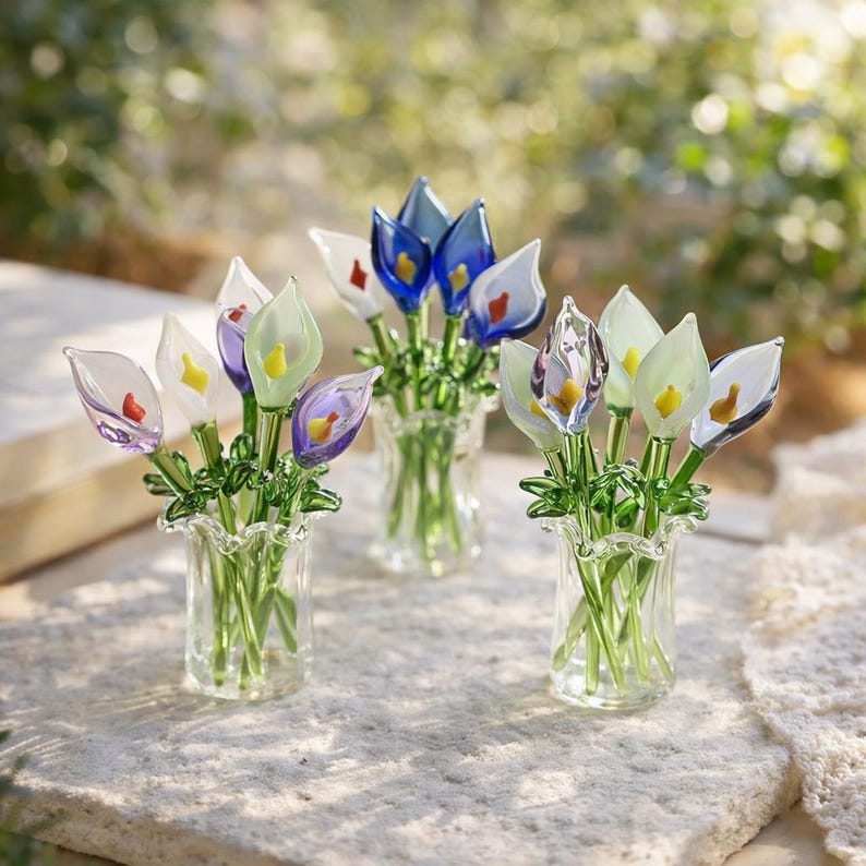 May include: Several small glass vases filled with colorful glass flowers. The flowers are in shades of white, blue, purple, and green, with yellow and red accents. The vases are clear and sit on a textured surface.