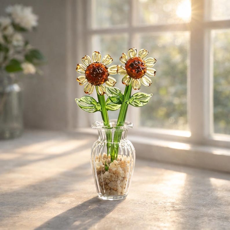 Puede incluir: Un jarr&oacute;n de cristal transparente con dos girasoles de cristal. Los girasoles tienen p&eacute;talos amarillos y centros marrones. Los tallos y las hojas son verdes. El jarr&oacute;n est&aacute; lleno de peque&ntilde;as piedras beige. El arreglo est&aacute; sobre una superficie clara.