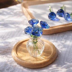 May include: A clear glass vase with a small bouquet of blue glass roses, set on a wooden tray. The roses have detailed petals and are arranged in a small, clear glass vase. Additional blue glass roses are visible in the background.