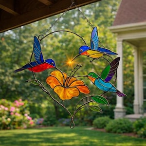 K&ouml;nnte beinhalten: Buntglas-Fensterbild mit Kolibris und einer Blume. Das farbenfrohe Design zeigt drei Kolibris mit blauen, roten und gr&uuml;nen Fl&uuml;geln und eine gelb-orangefarbene Blume. Es h&auml;ngt an einer Kette.