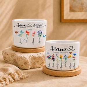 May include: Two white ceramic plant pots with floral designs and personalised text. One reads "James & Sarah Brown 2026" with names and flowers. The other says "Nana Growing Since 2025" with names and flowers. Both sit on wooden coasters.