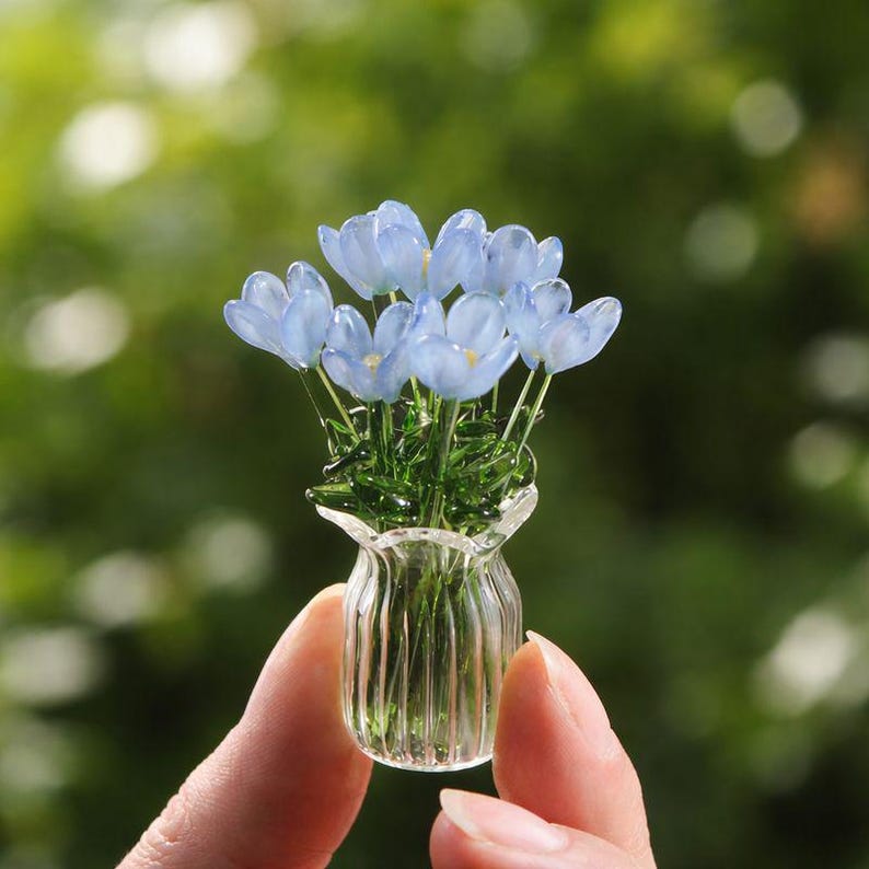 May include: A miniature glass vase filled with delicate blue flowers. The vase has a fluted design and is held between fingers against a blurred green background. The flowers have translucent petals and yellow centers, creating a whimsical floral arrangement.