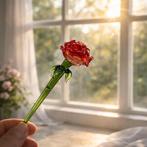 May include: A handcrafted glass rose with a vibrant red bloom and green stem and leaves. The glass rose is held in front of a window with natural light.