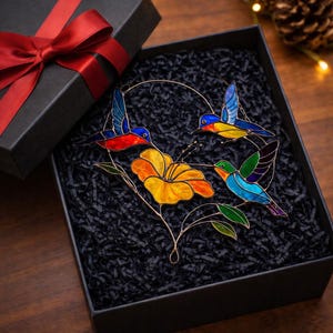 K&ouml;nnte beinhalten: Ein Buntglas-Kunstwerk mit Kolibris und Blumen in einer schwarzen Geschenkbox mit rotem Band. Das farbenfrohe Design zeigt drei Kolibris mit blauen, roten und gelben Fl&uuml;geln und eine gelb-orangefarbene Blume.