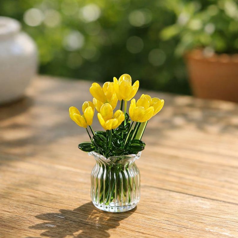 May include: A small glass vase with a fluted design holds a bouquet of yellow tulips. The tulips have green stems and leaves. The vase sits on a wooden surface, with a blurred green background.