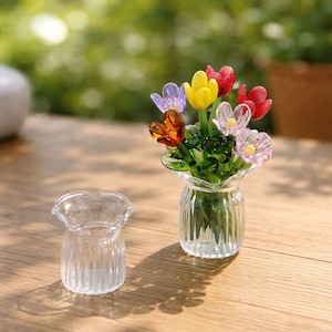 May include: A small glass vase filled with colorful glass tulip flowers. The tulips are various colors, including red, yellow, orange, and purple. A second, empty glass vase sits next to the vase of flowers. The vases are on a wooden surface.