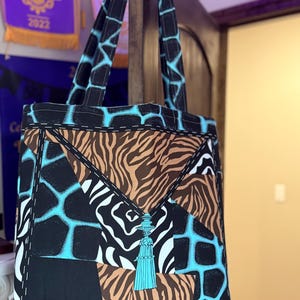 以下が含まれることがあります： 黒、茶色、白の様々なアニマルプリント柄を組み合わせたパッチワークトートバッグ。 ターコイズブルーのアクセントが施されています。 ターコイズブルーの幾何学模様と装飾的なタッセルが付いた黒いハンドルが付いています。