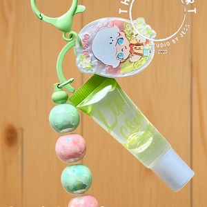 May include: A beaded lipgloss keychain with a light green clasp, a charm with a cartoon character, and a tube of lip gloss. The keychain has a string of green and pink beads. The text "Beaded Lipgloss Keychain!" is at the bottom.