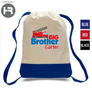 May include: A beige drawstring backpack with a blue bottom panel and blue shoulder cords. The front features a red fire truck graphic above the blue text "BIG Brother" and the red text "Carter" printed in a bold, sans-serif font style.