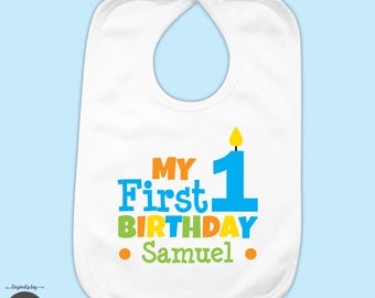 May include: A white baby bib featuring the text "MY First 1 BIRTHDAY Samuel" in blue, orange, and green lettering. The number one is designed as a blue candle with a yellow flame. Two orange dots flank the name Samuel at the bottom of the bib.