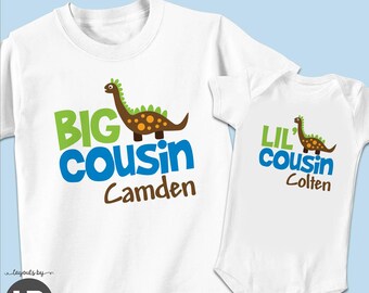 May include: A white t-shirt and a white baby bodysuit set featuring a brown dinosaur graphic. The t-shirt displays the text BIG COUSIN Camden in green, blue, and brown. The bodysuit displays the text LIL' COUSIN Colten. LONG OR SHORT SLEEVE * SET OF 2.
