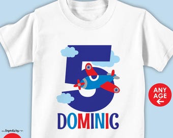 May include: White t-shirt with a blue number 5 and a red and blue airplane flying over it. The name DOMINIC is written below the number. The shirt is available in short or long sleeve and shirt or bodysuit.